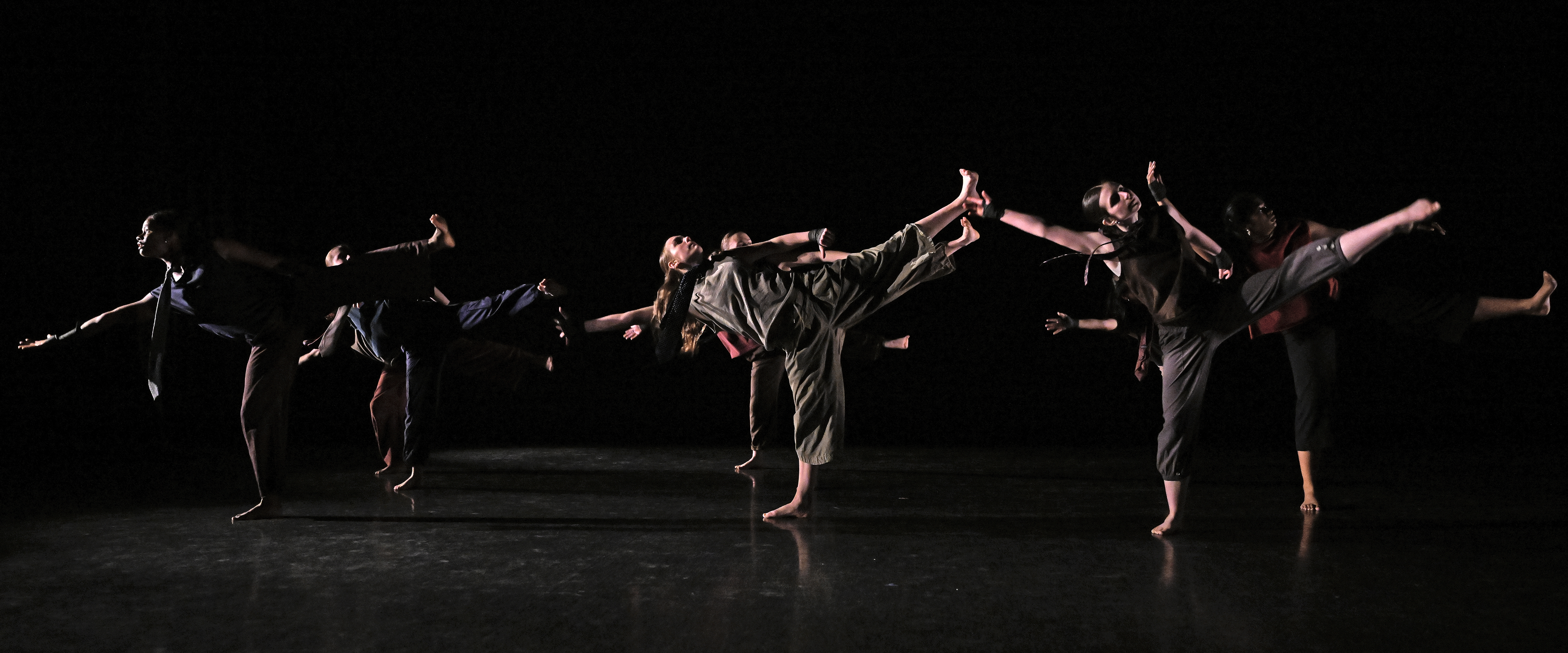 Student dancers with one leg raised in the air perform in a dark dance studio.