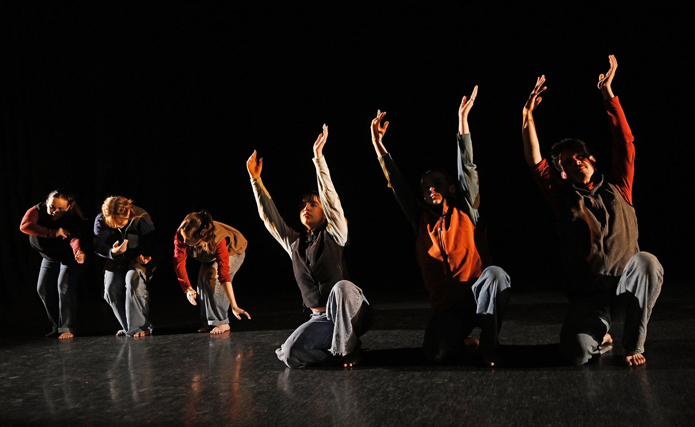 Student Dancers, three of them kneeling with arms raised, all dressed in jeans and vests, perform in a dark dance studio.