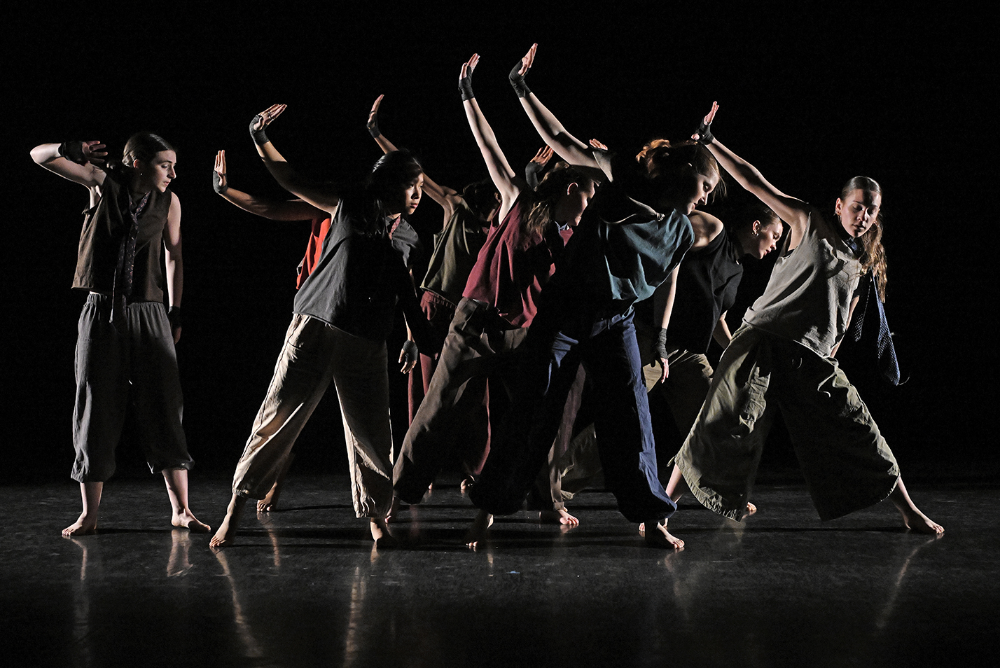 A group of student dancers in varied-colored outfits reach high with one arm while performing in a dark dance studio.