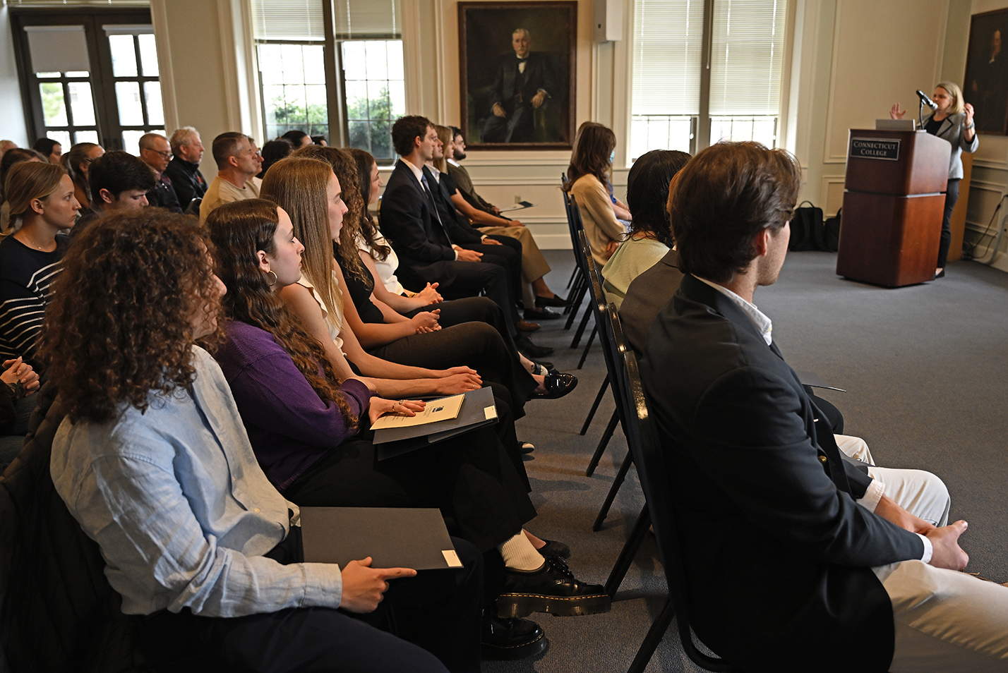 Seated students listen as their college president addresses them from a podium at the front of a conference room during an honor society induction ceremony.