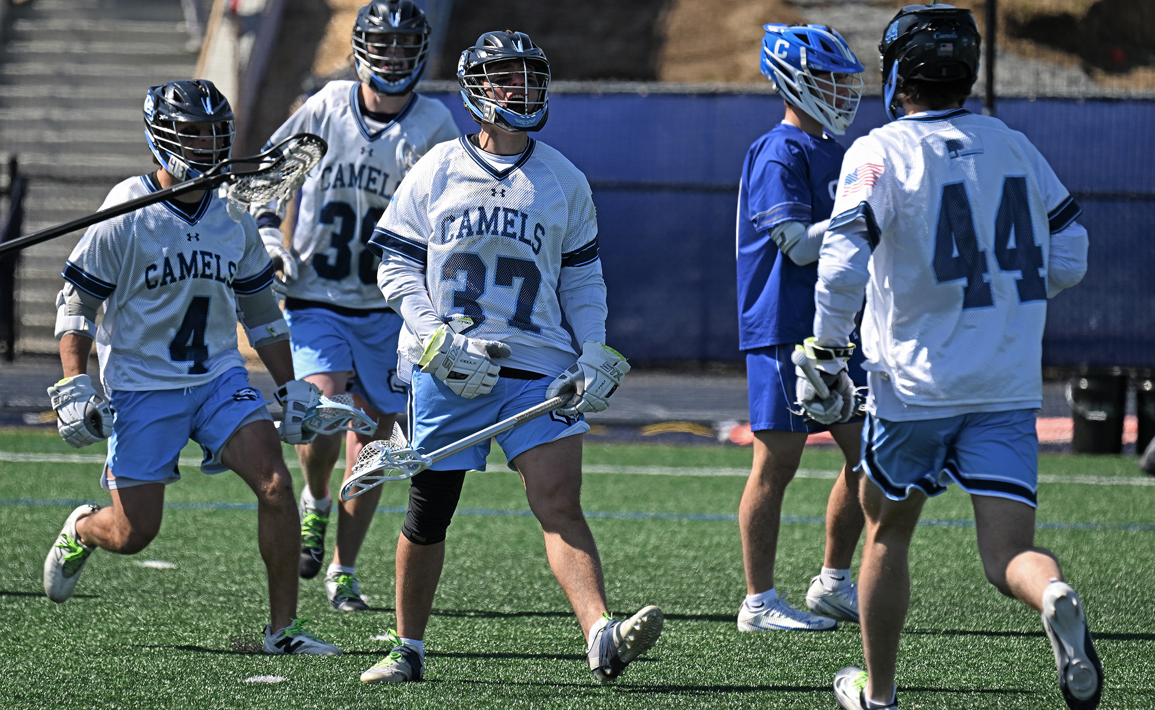 A men's lacrosse player lets out a yell as teammates flock to him following a goal on a turf field.