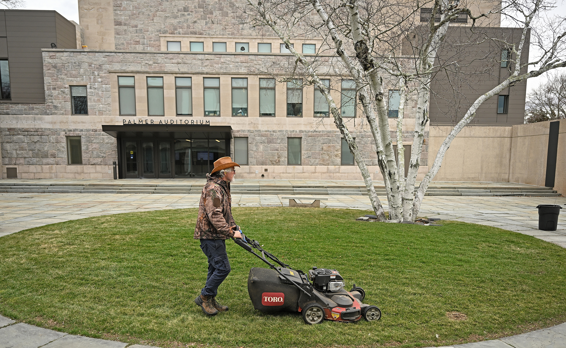 A college grounds crew members in camofluage jacket and cowboy hat uses a push lawnmower to trip the grass in a small green in a courtyard.