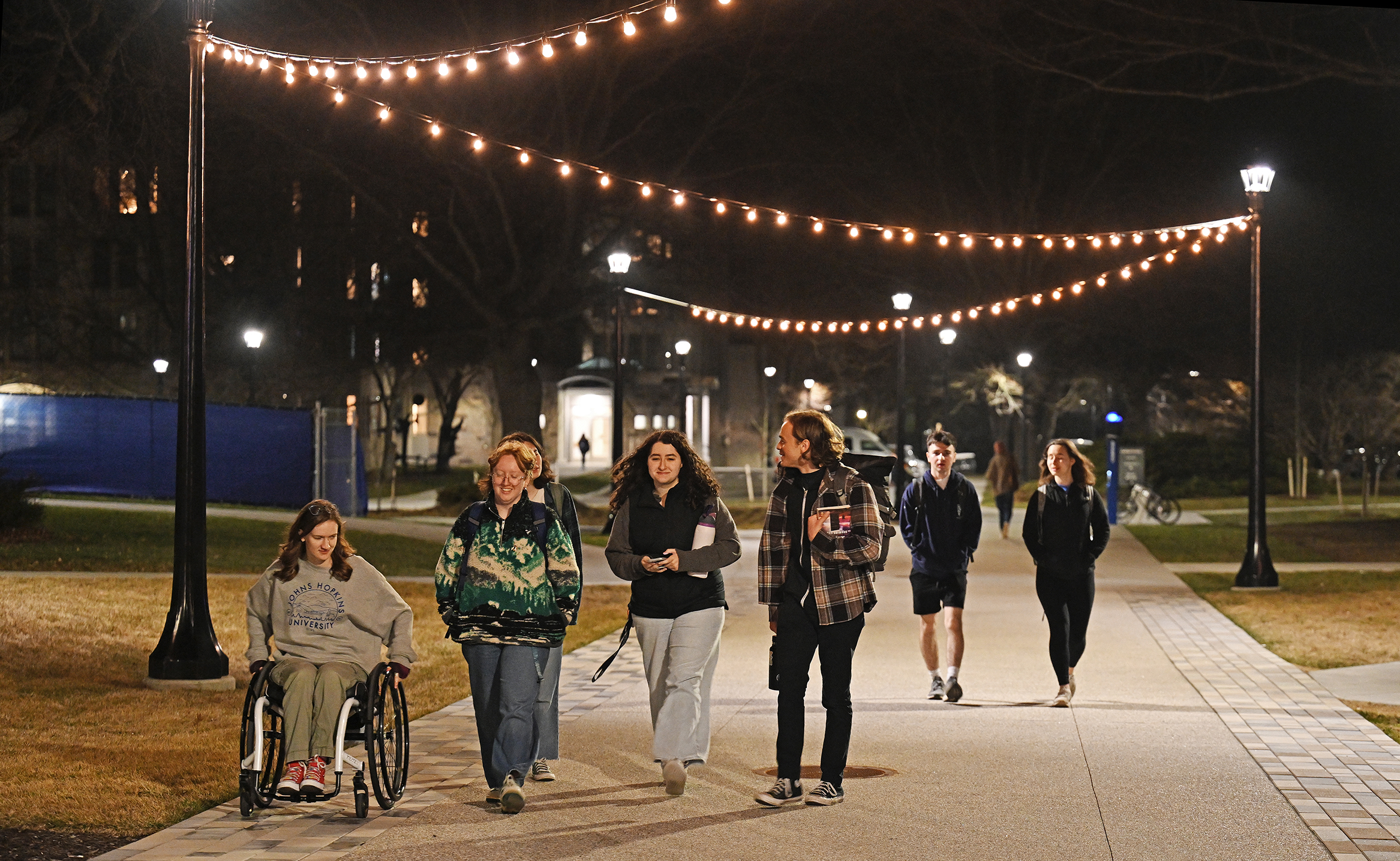 College students, one in a wheelchair, all wearing jackets and carrying boos and water bottles, walk along a pedestrian path with small lights strung overhead between lampposts.