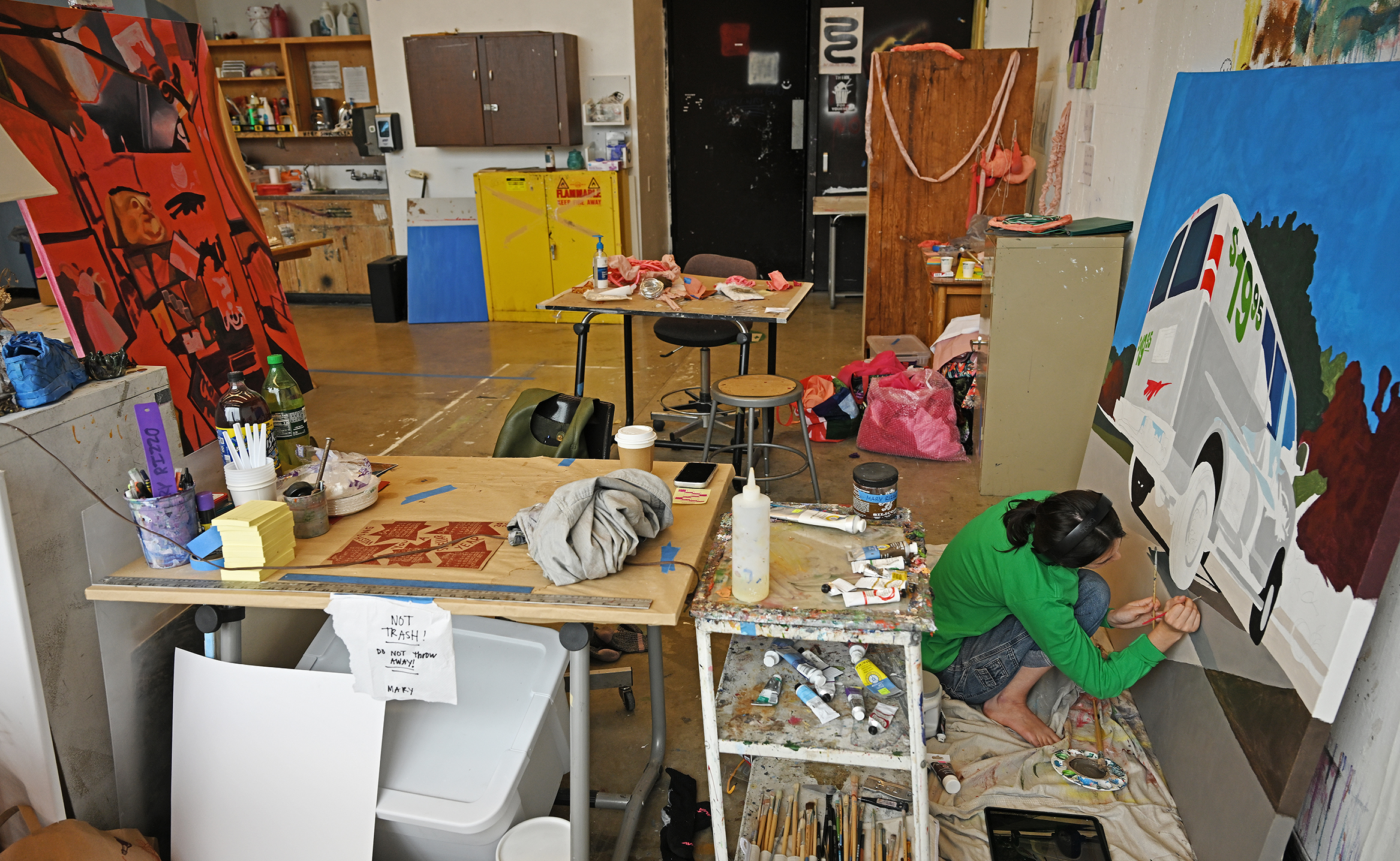 A student artist in green long-sleeve t-shirt paints a large format painting depicting a U-Haul van in an art studio setting.