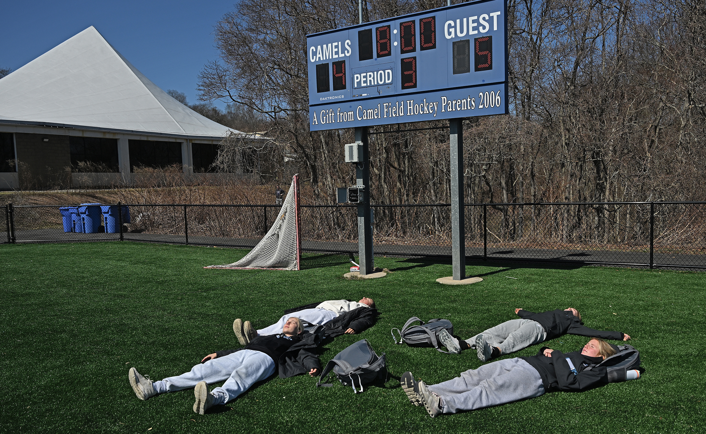 A group of female college athletes in their workout clothes recline on a patch of artificial turf under a scoreboard showing a 5-4 lead for the visiting team.