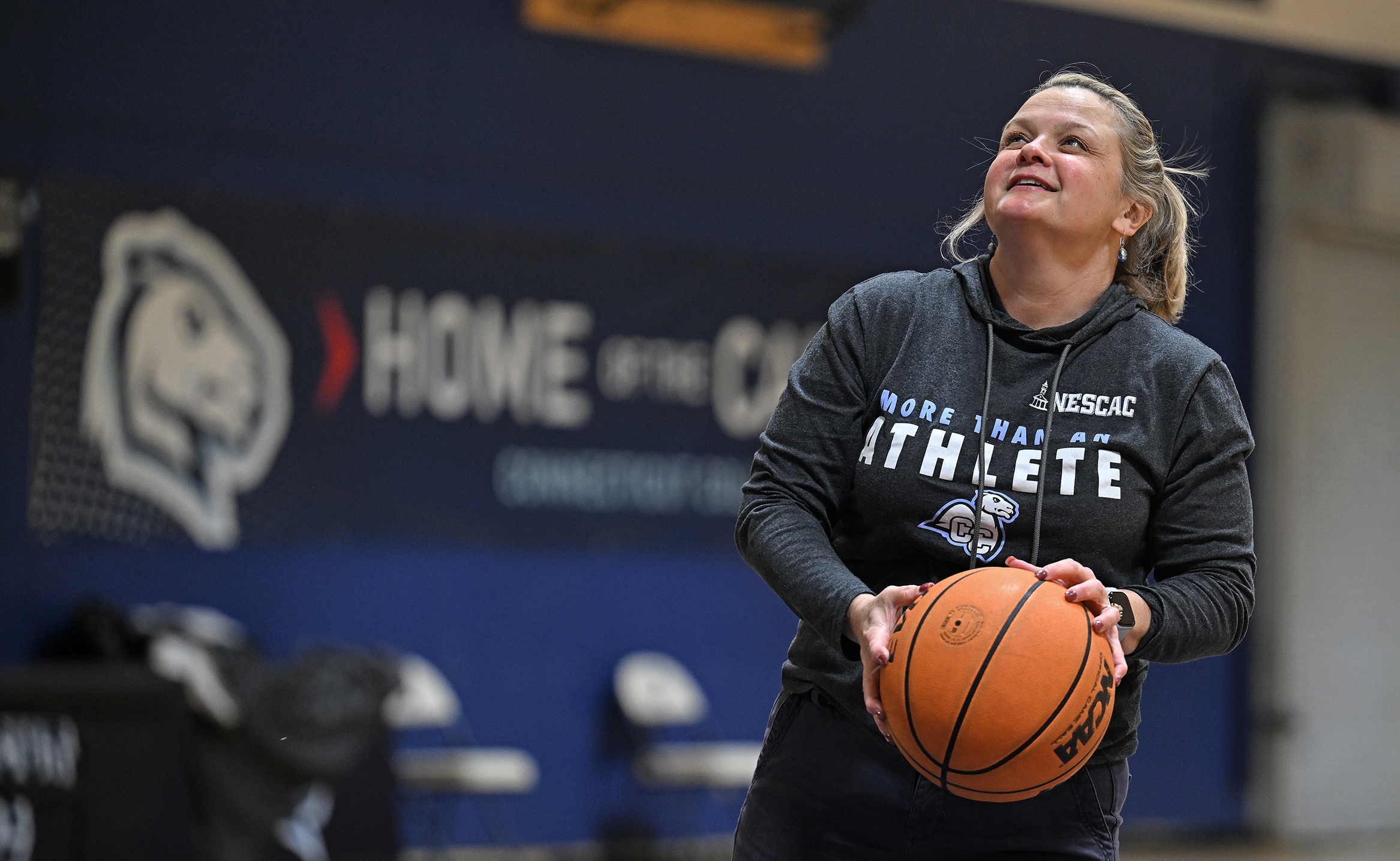 A college president in athletic swag hoodie prepares to take a jump shot on a college basketball court.
