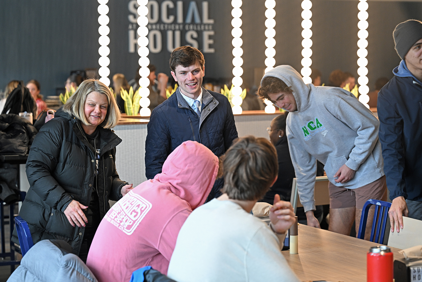 A college president in winter coat prepares to join a group of student athletes for lunch in a college dining hall.