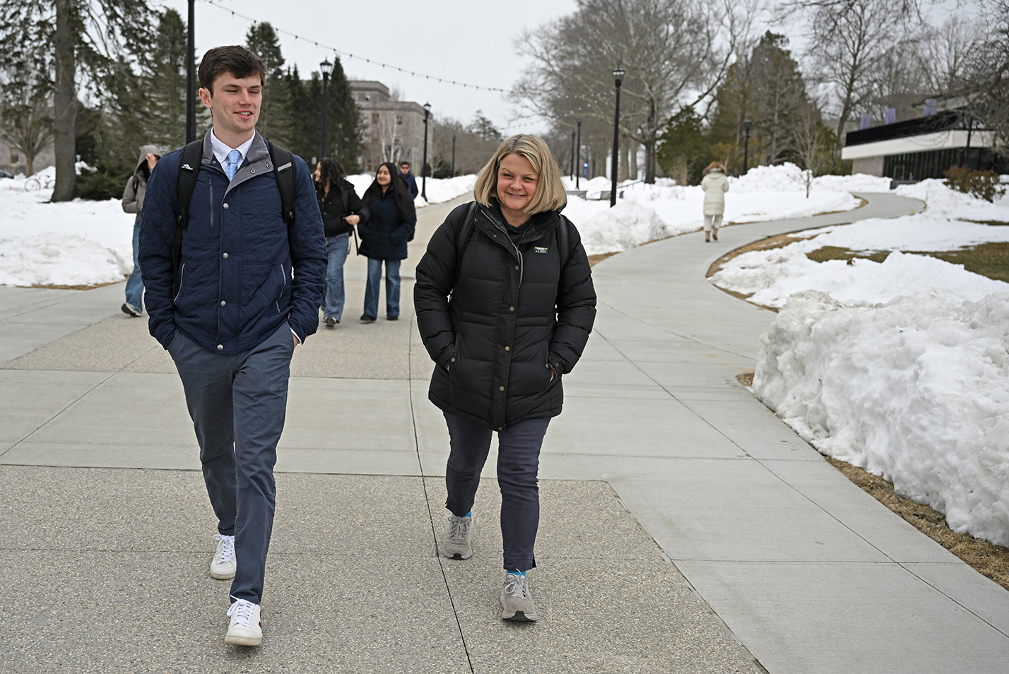 A college president in a winter coat walks with a student athlete in shirt and tie and coat along a snow-flanked pedestrian path.