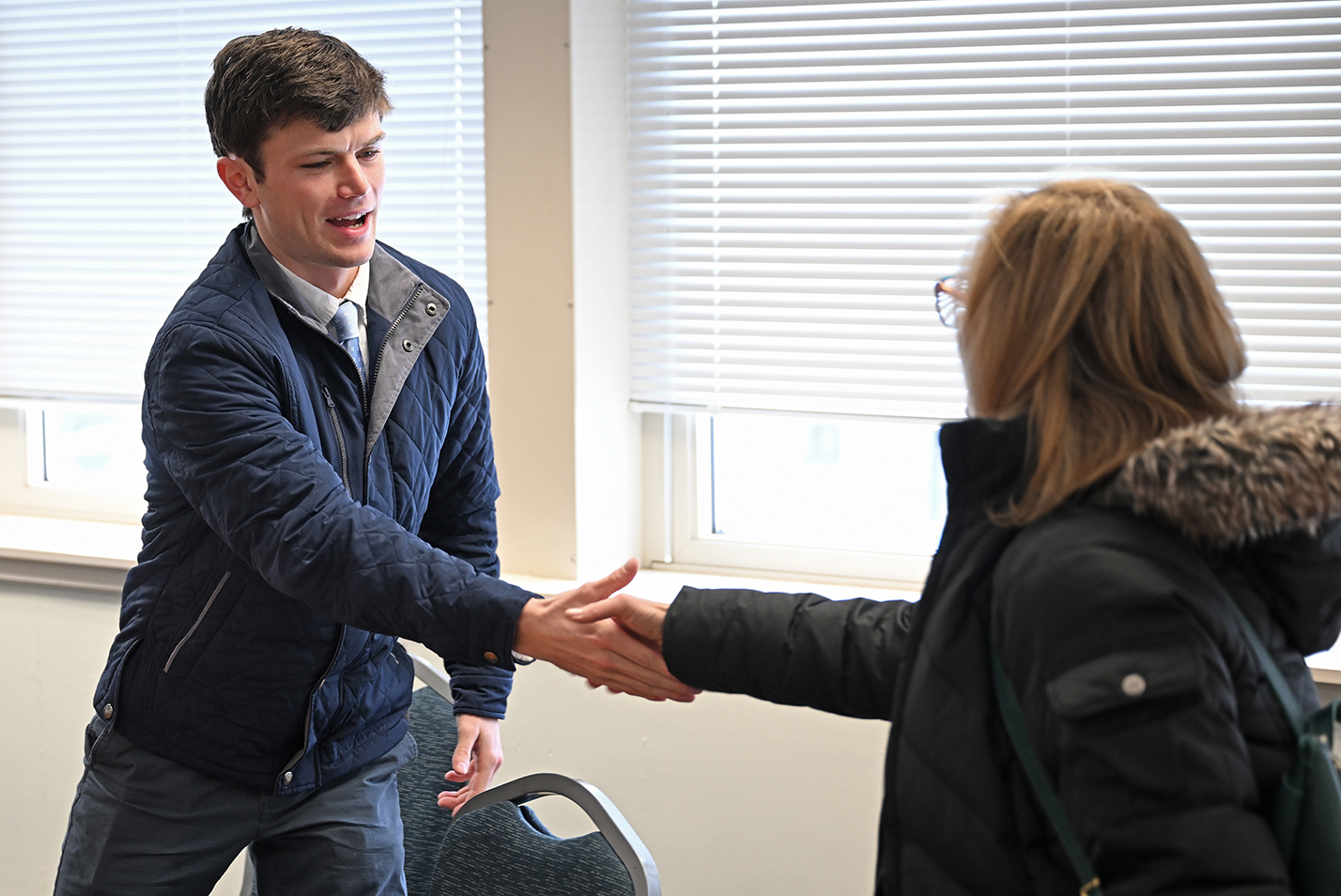 A college student athlete in winter jacket and shirt and tie reaches to shake hands with a college administrator during a meeting in a conference room.