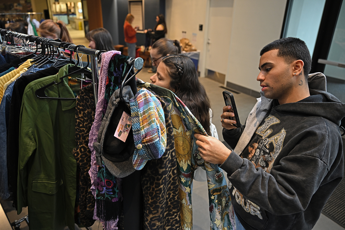 A college student uses his smart phone to show a friend some thrift clothing on a rack during a thrift shop in a college student center.