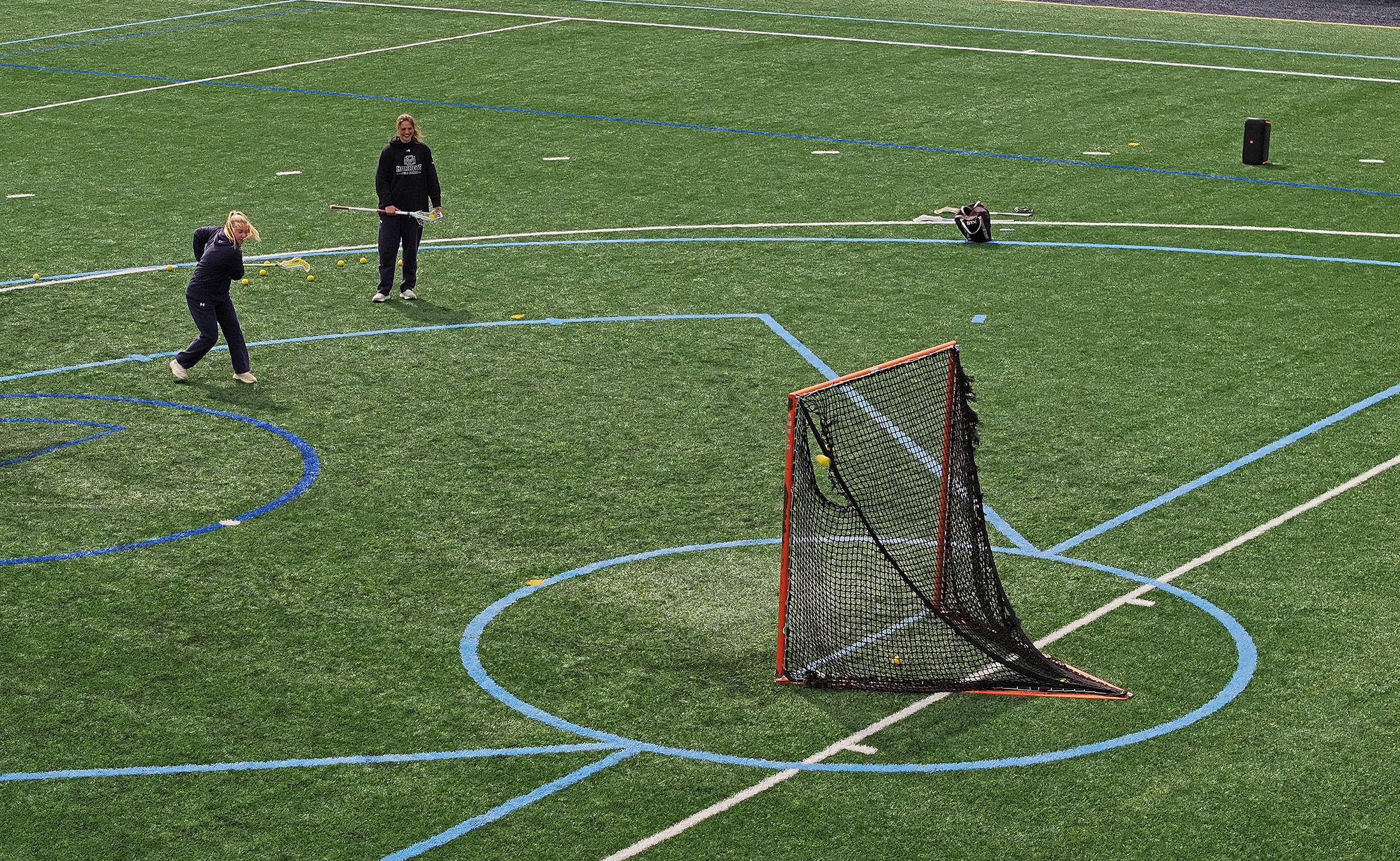 Two women's lacrosse players, seen from above, take practice shots at a net on an artificial turf field.