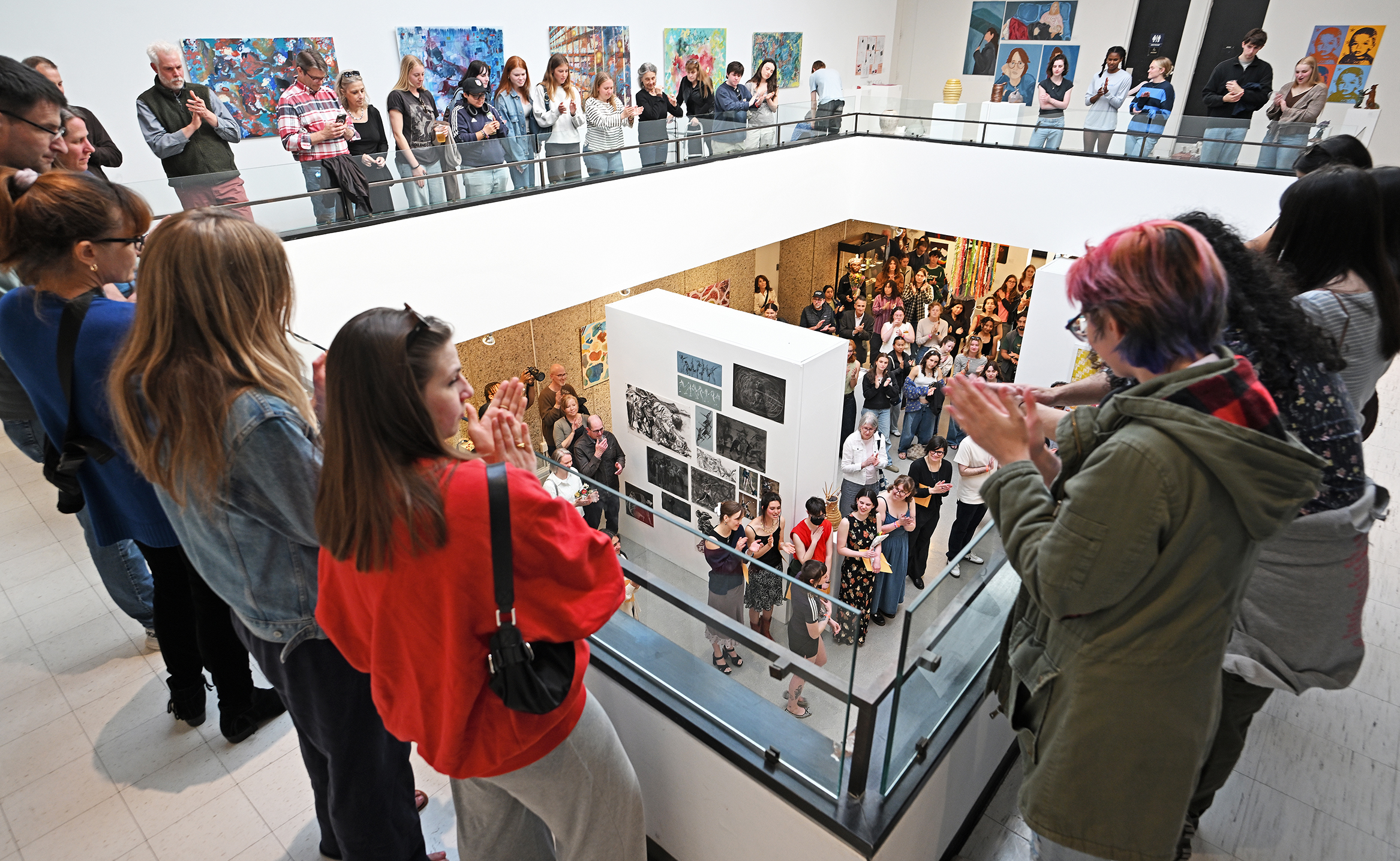 A crowd lines a balcony level around an art gallery applauding the student artists gathered below.