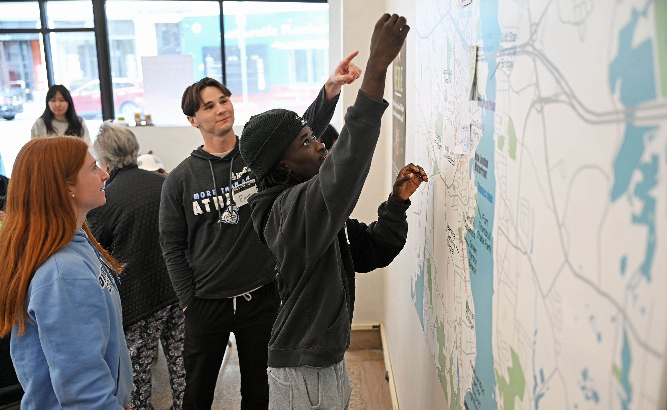 Three college students, one female with red hair, one black wearing a cap, and one white wearing an athletics sweatshirt, place small notes on a giant wall map.