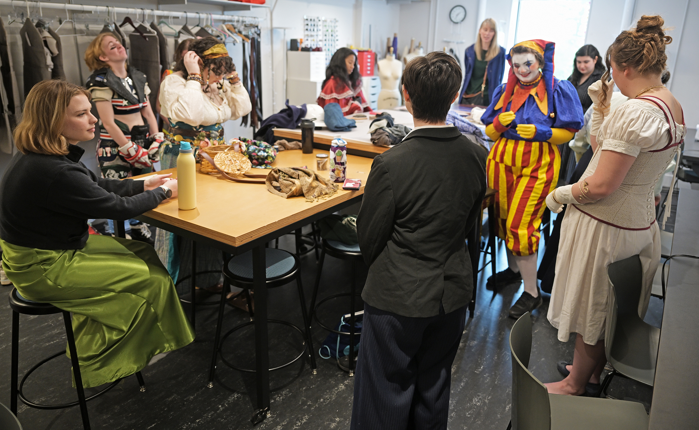 A group of college theater students in a variety of costumes, including one in bright red and yellow clown outfit, gather around the work table in a costume shop to present final projects in a costuming course.