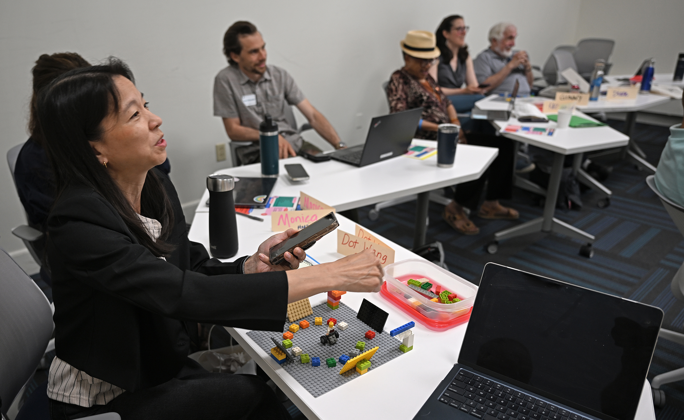 A workshop participant presents a model classroom made out of LEGO bricks to a group of attendees seated at seminar room tables.