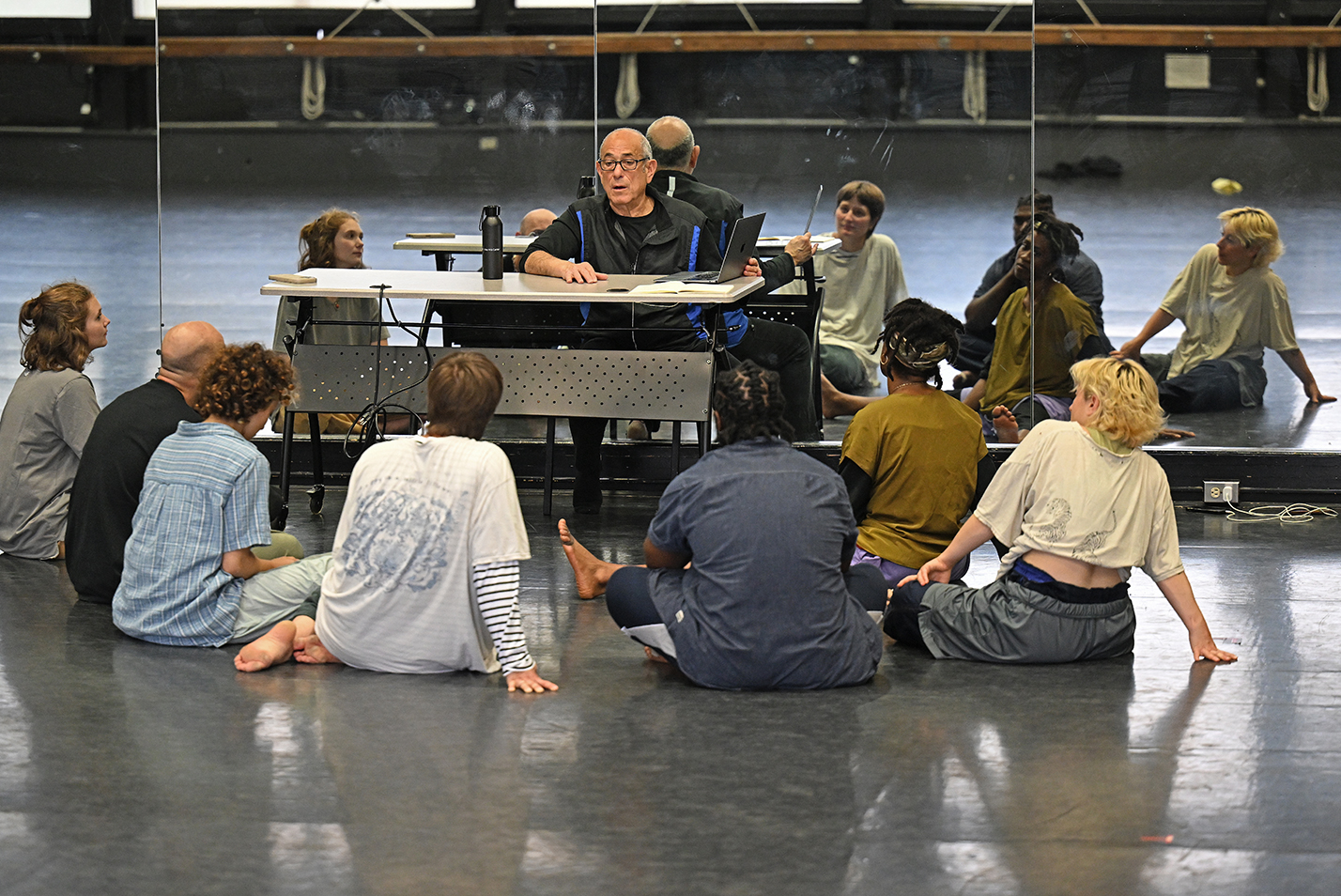 A choreographer sits at a table and goes over notes with a group of modern dancers all seated on the floor in front of a dance studio mirrored wall.