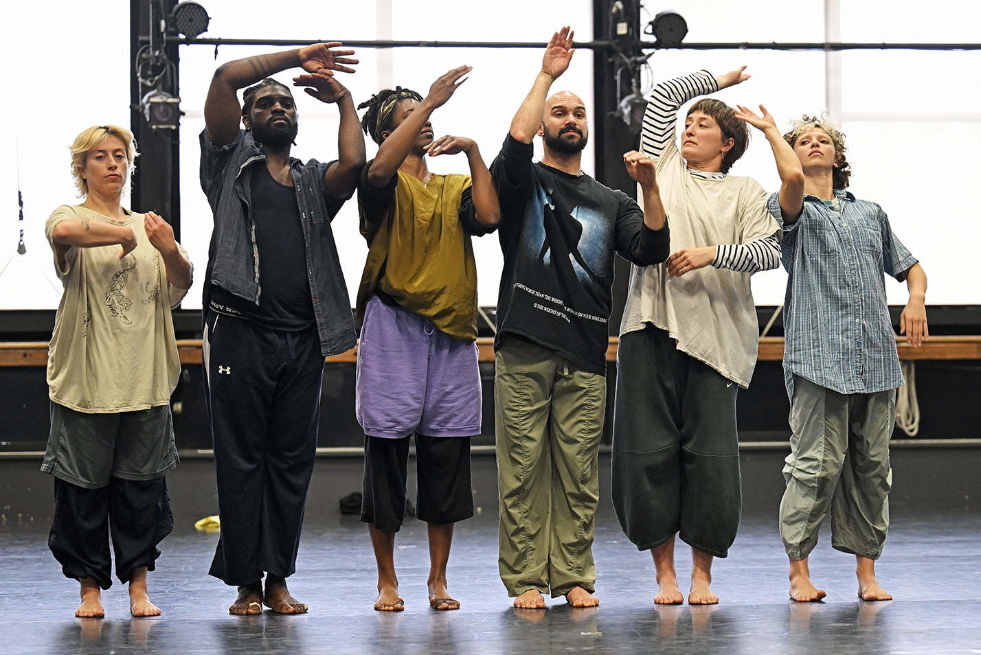 Seven members of a modern dance company stand in a line and gesture wildly with their arms in a dance studio.