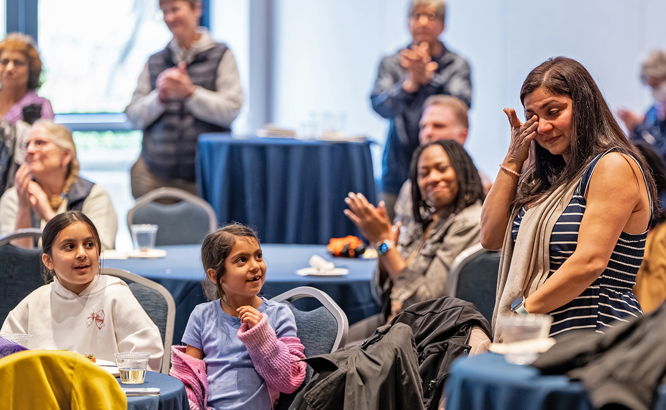 A female college professor in a horizontally white striped tank dress wipes a tear as fellow faculty members and her two children look on and applaud during an awards ceremony.