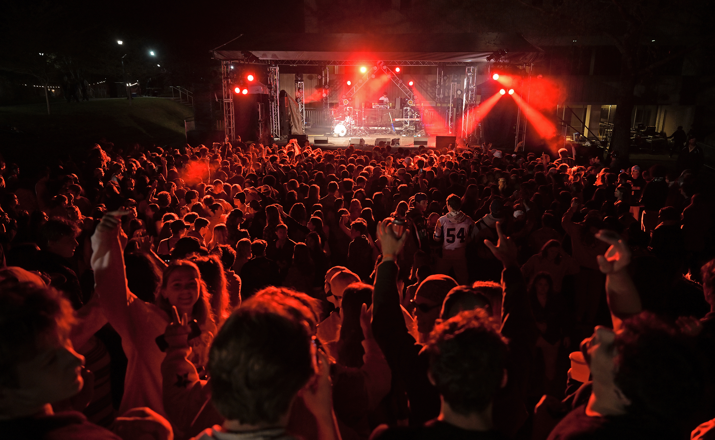 A crowd of college students bathed in red light fill a small outdoor music venue as a DJ performs on stage.