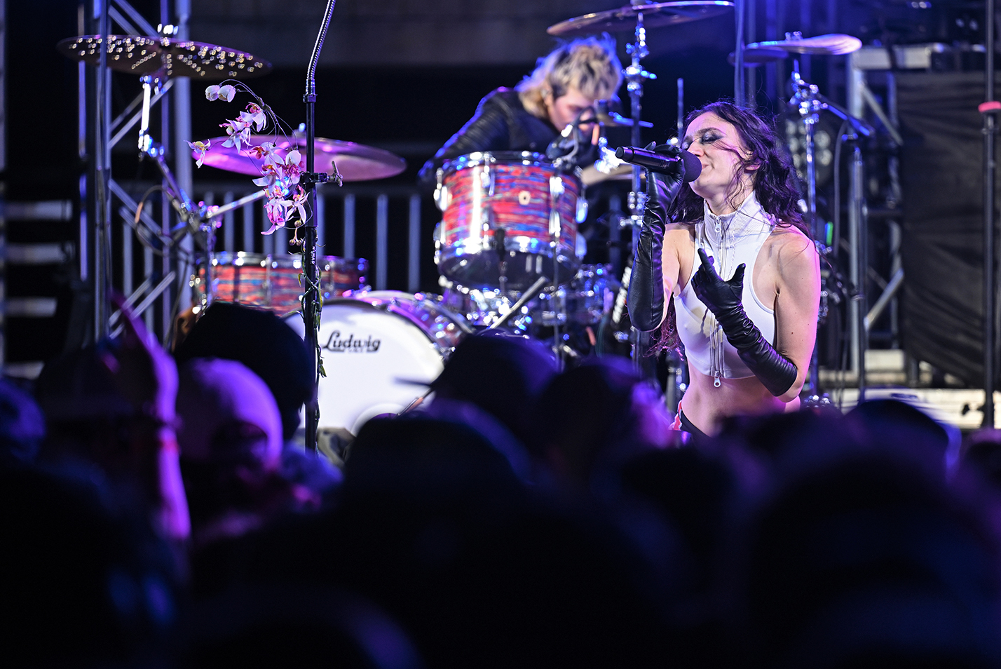 A pop singer in turtle necked tank top and long black gloves kneels on stage in front of a crowd as a drummer plays in the background.