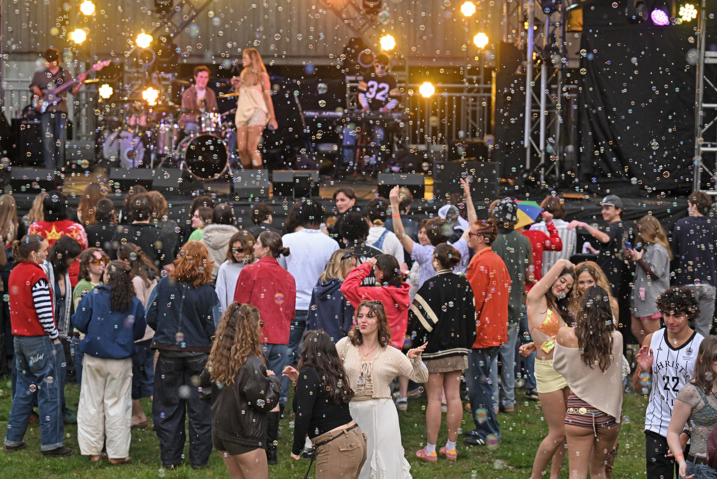 A cloud of small soap bubbles fills the air as college students dance on the grass to a student band at an outdoor concert.
