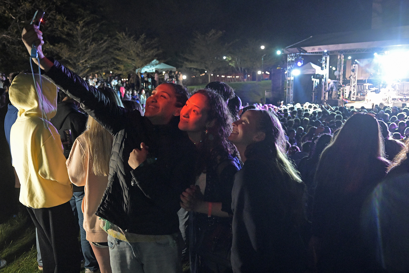 Three female college students at the edge of a large nighttime concert crowd take a photo together illuminated by the orange glow from the camera.