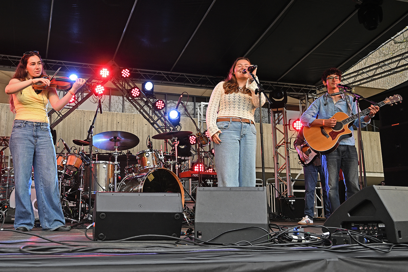 Members of a student bluegrass band, one on violin, one on acoustic guitar, one singing vocals, perform on an outdoor stage.