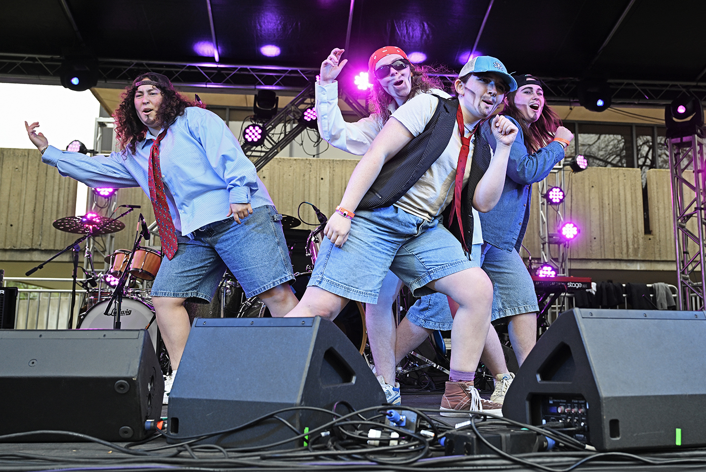 A quartet of female-presenting students in boy band drag and painted-on facial hair lip syncs on stage at an outdoor concert.