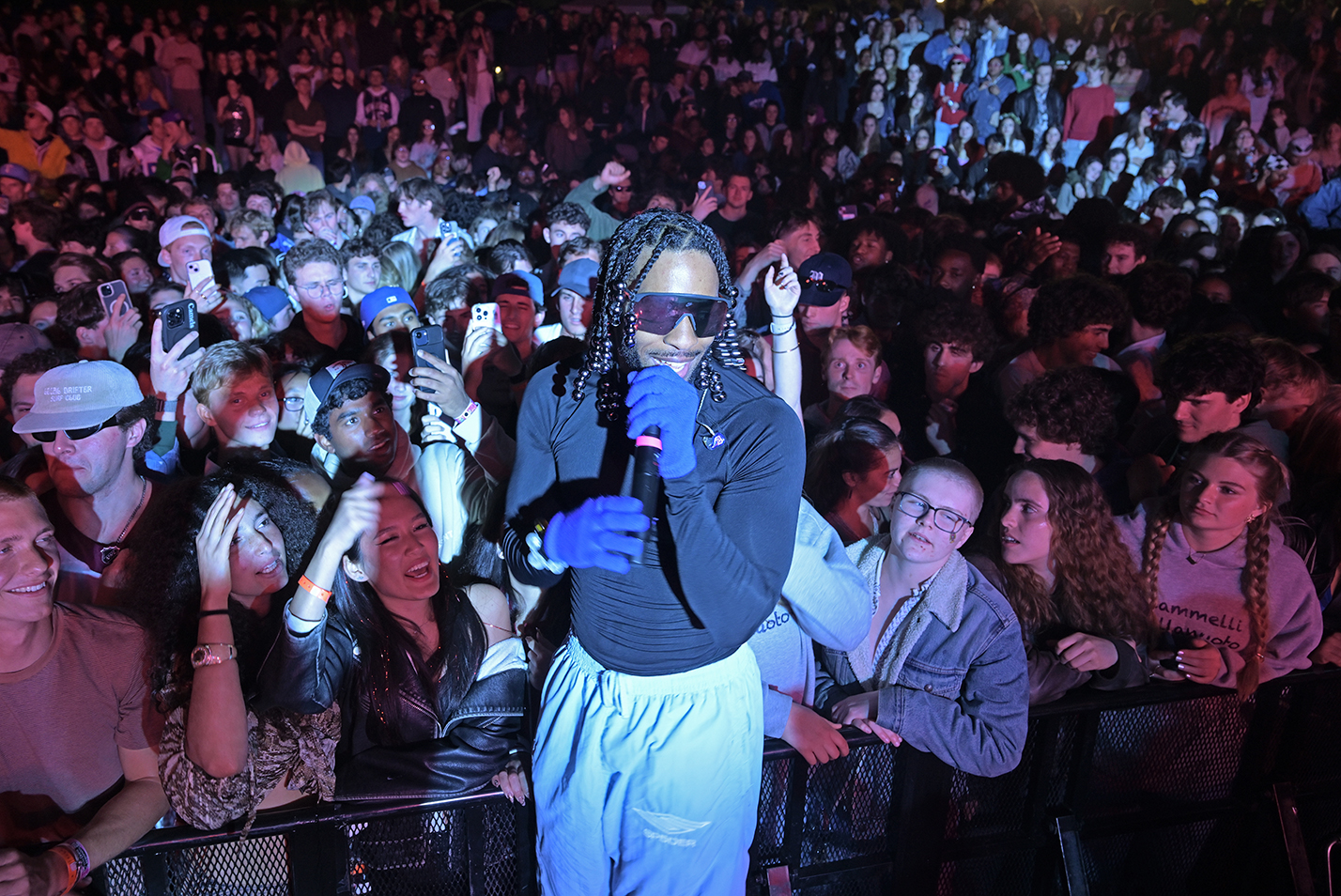 A black male rapper in long sleeved shirt and sweatpants and illuminated by a spotlight stands elevated in front of the audience of college students smiling, and cheering, and taking photos.