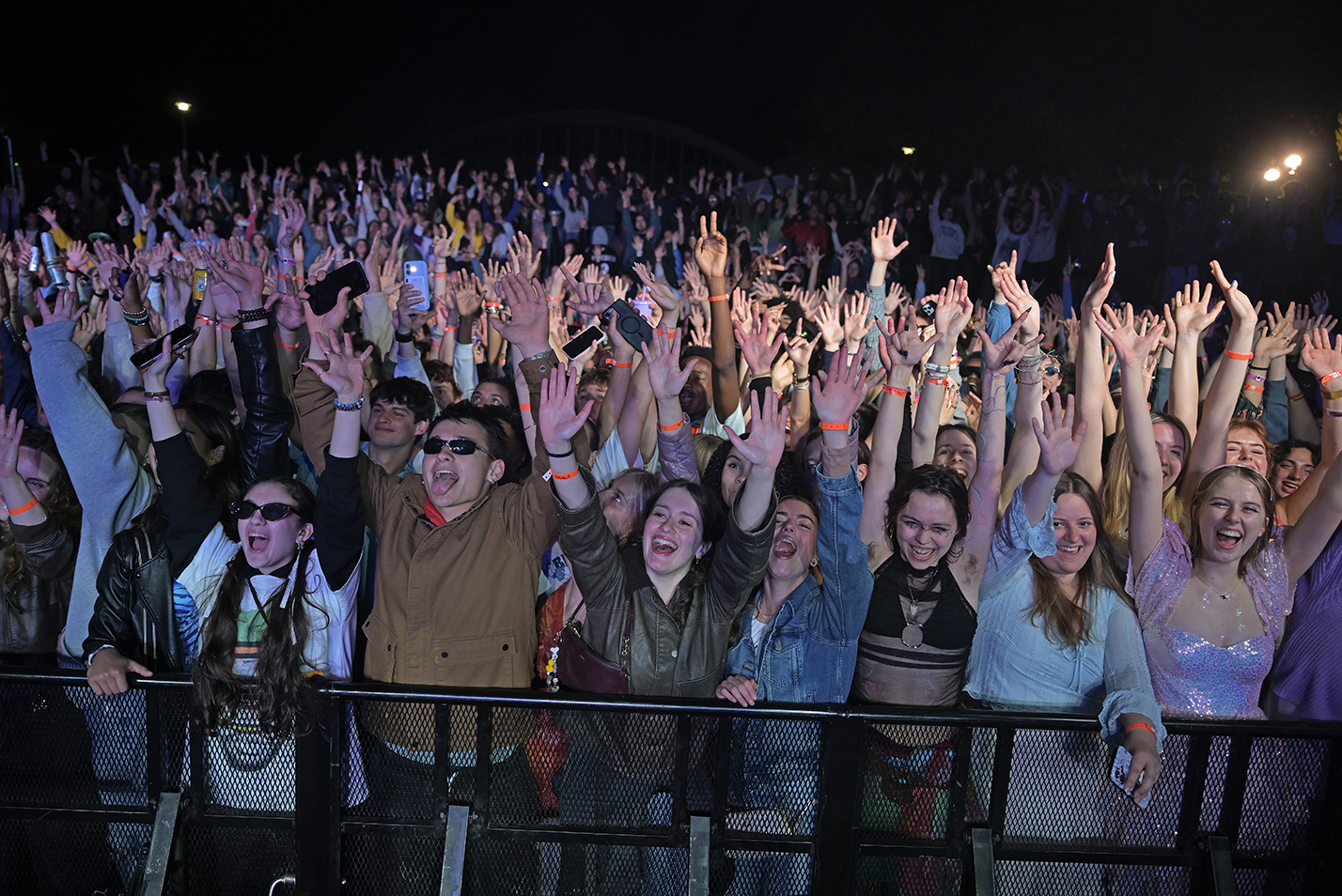 A large crowd of college students hold their hands in the air as they cheer a performance on stage at an outdoor concert.