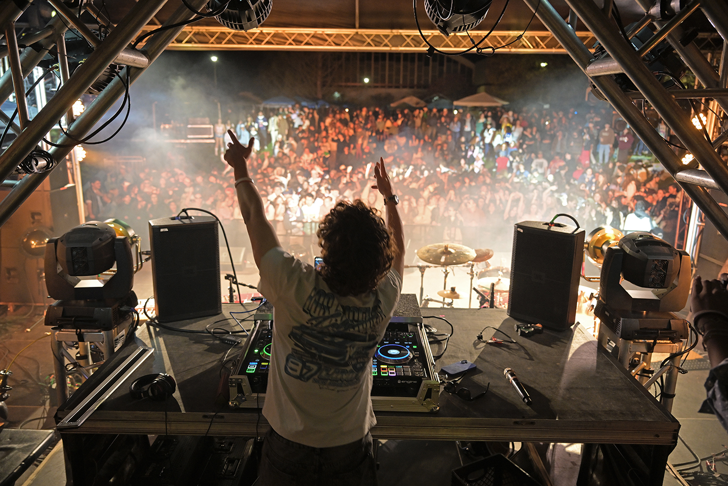 A DJ at his mixing board holds his hands in the air to the audience in the distance during a nighttime outdoor concert.