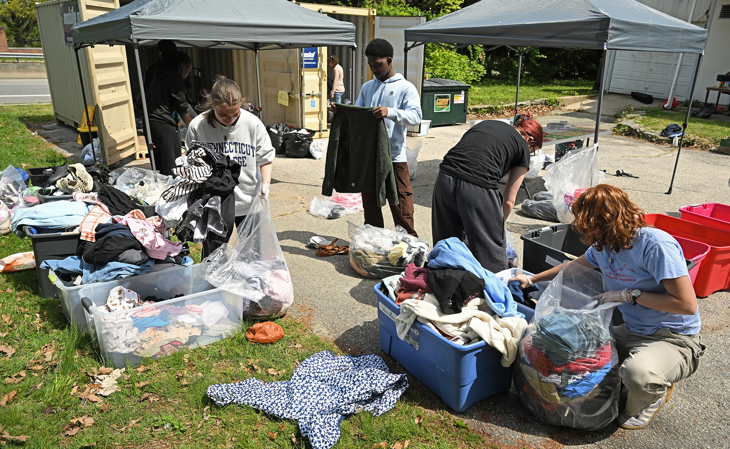 College students sort through clothing donations sitting on the lawn or standing in the driveway of a campus building with a portable tent and a garage and storage unit in the background.