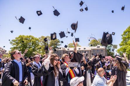 Graduation caps fly in the air tossed by the graduates at a college commencement ceremony under a clear blue sky.