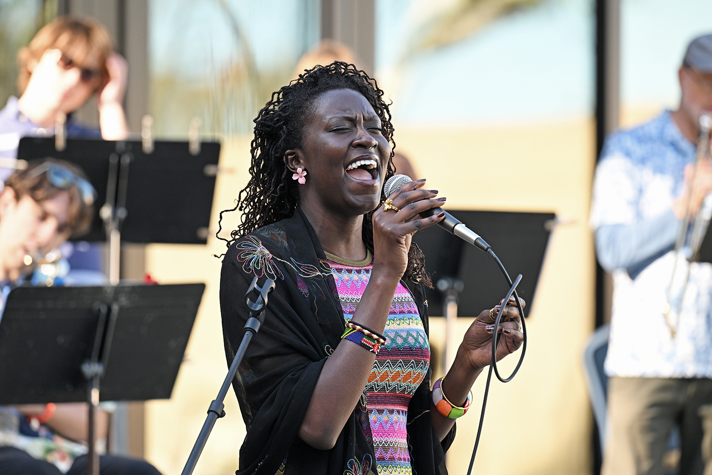A black female college student in a brightly colored and patterned dress, with black jacket, sings expressively in front of a jazz band at an outdoor courtyard.