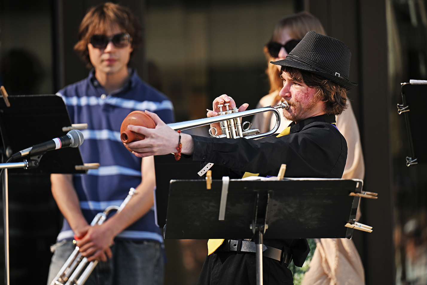 A male student musician in black shirt and black fedora hat plays the trumpet using a sink plunger head as a mute at an outdoor concert.