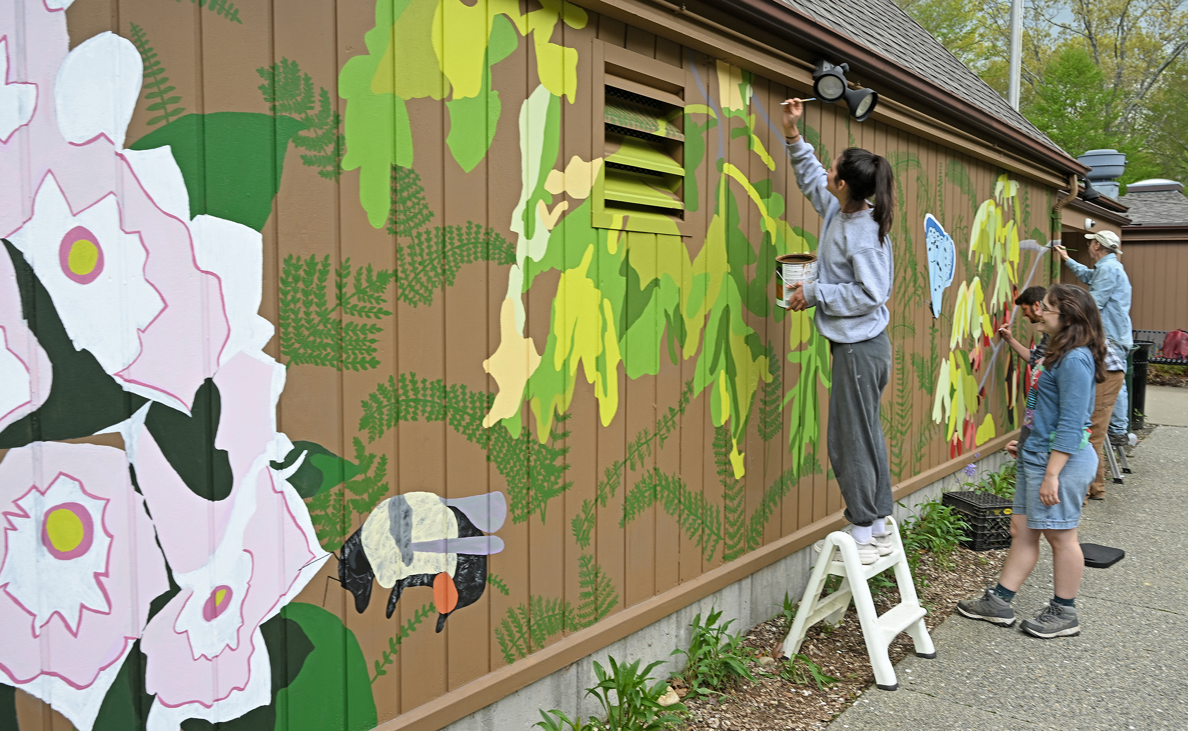 A student artist on a short stepladder paints a detail on a large wall mural as other helpers work on other parts of the mural, depicting flowers and plants and insects.