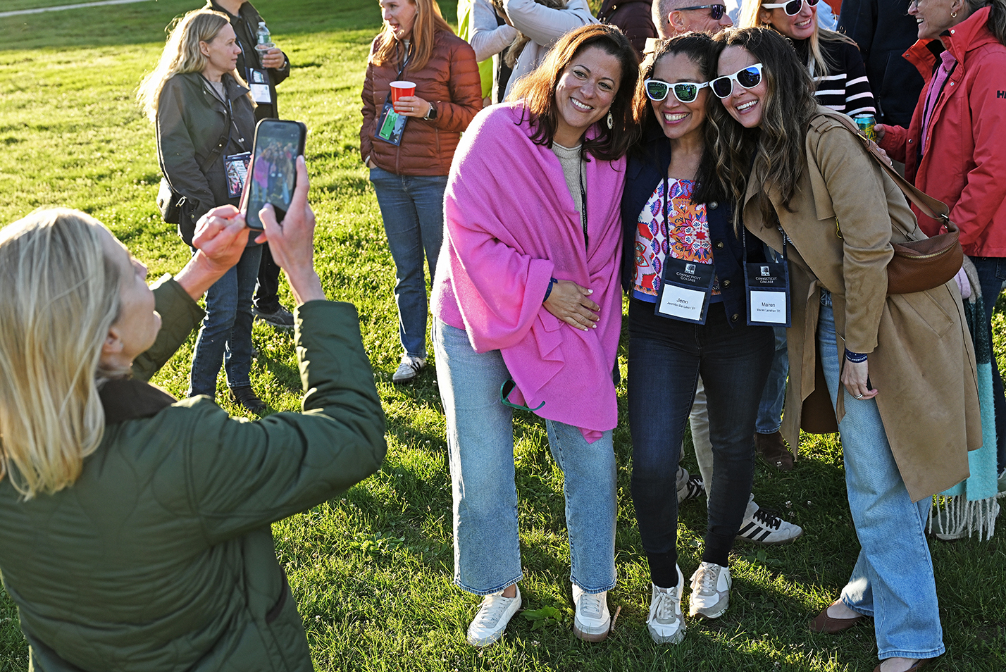 Three college alumni in sunglasses and wearing name tags smile for a classmate with a smart phone camera in late afternoon light.