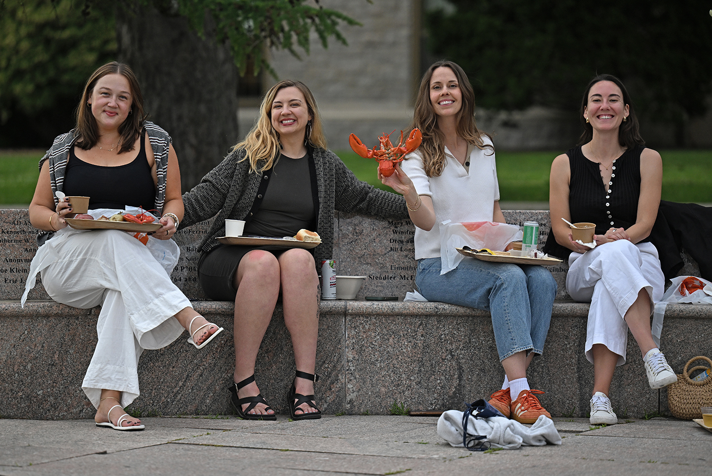 For female college alumni seated on a granite outdoor bench, one holding up a bright orange cooked lobster, smile for a photo.