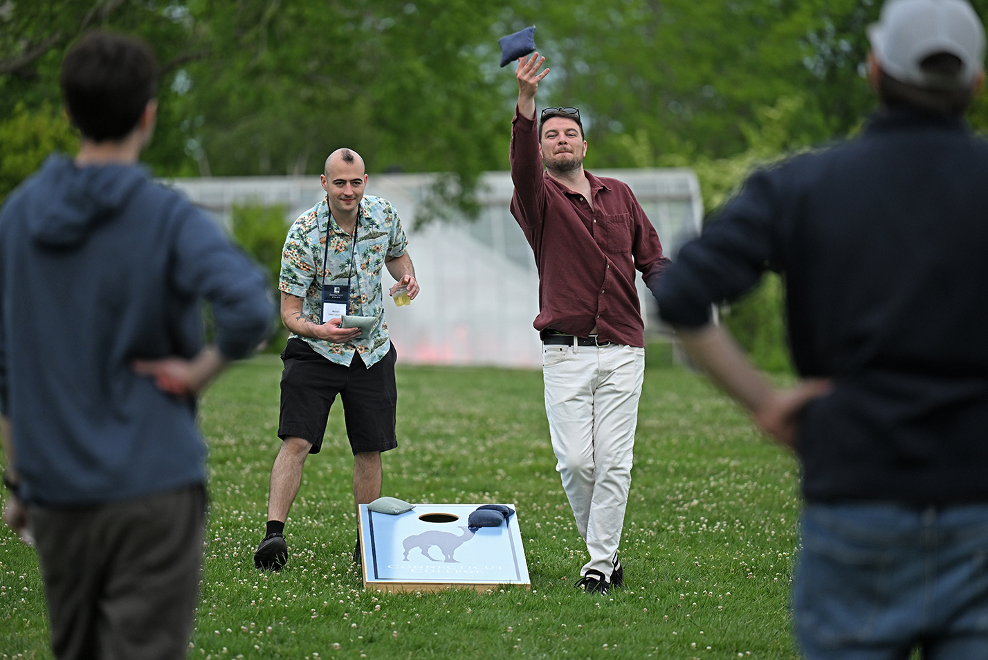 Four young adult college alumi, two facing the viewer, two facing away and framing the first two, play a game of Cornhole on a college green.