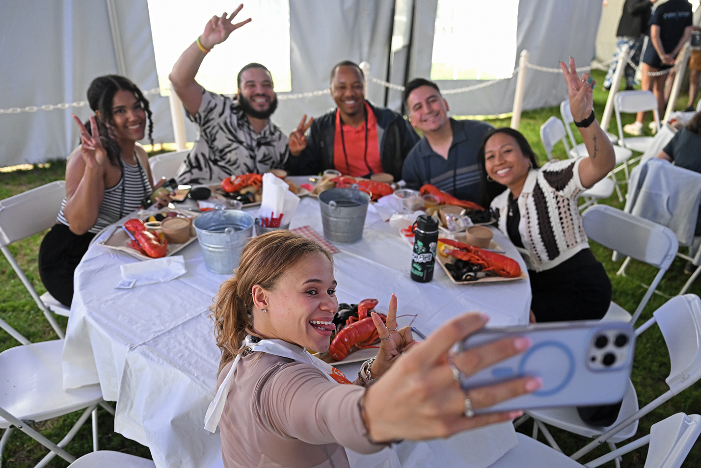 A college alumna holding a smart phone strikes a pose as classmates across a round dinner table with bright orange cooked lobsters on their plates smile and pose for the photo.