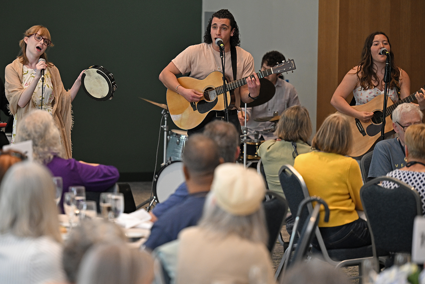 College student musicians, two with acoustic guitars and one with a tambourine perform for a luncheon gathering in a ballroom space.