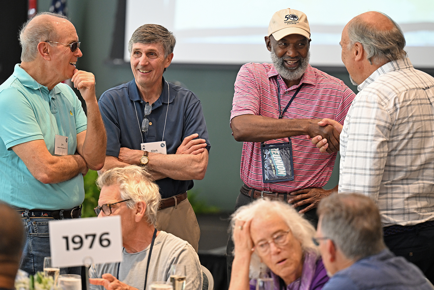 Four male college alumni from the class of 1976 stand and talk in a crowded ballroom.