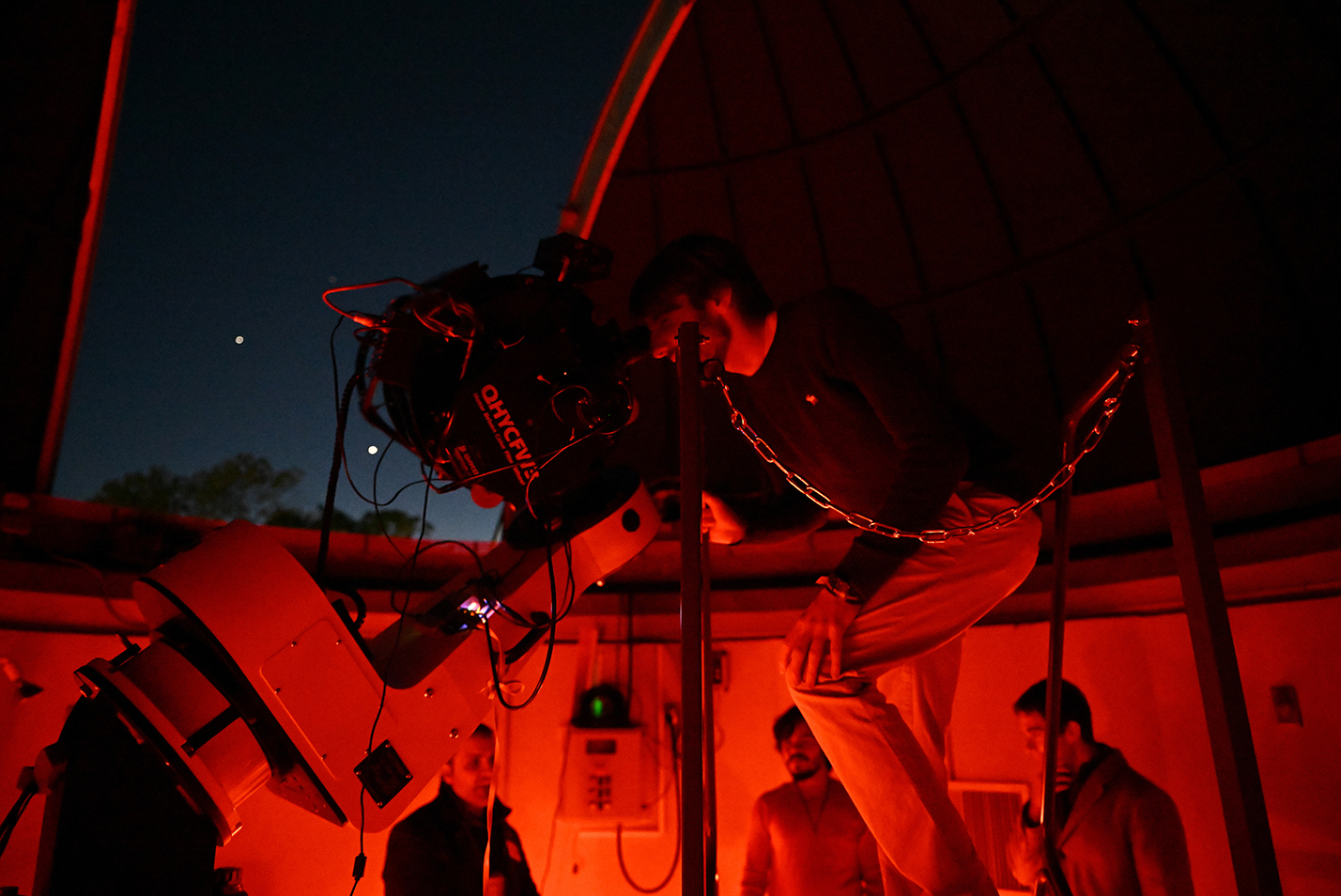 A person bathed in a red glow bends over to peer into a telescope pointed out the large open door of a rooftop observatory as bright planets glow in the dark sky beyond.