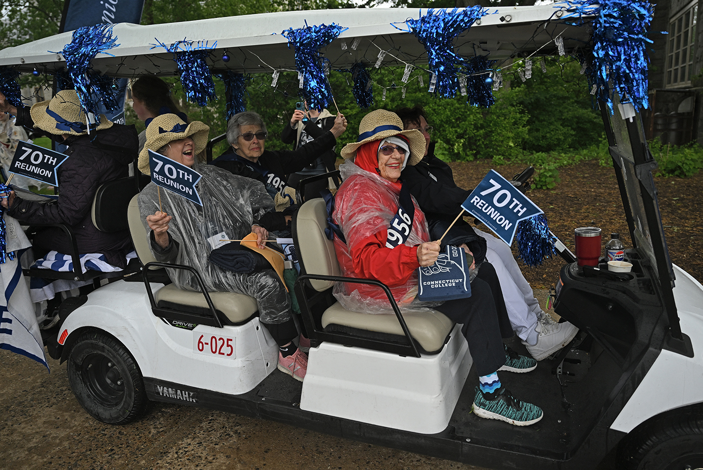 A group of college alumni, carrying 70th Reunion pennants, wearing broad brimmed hats and raincoats, ride in a golf cart decorated with blue pom poms.