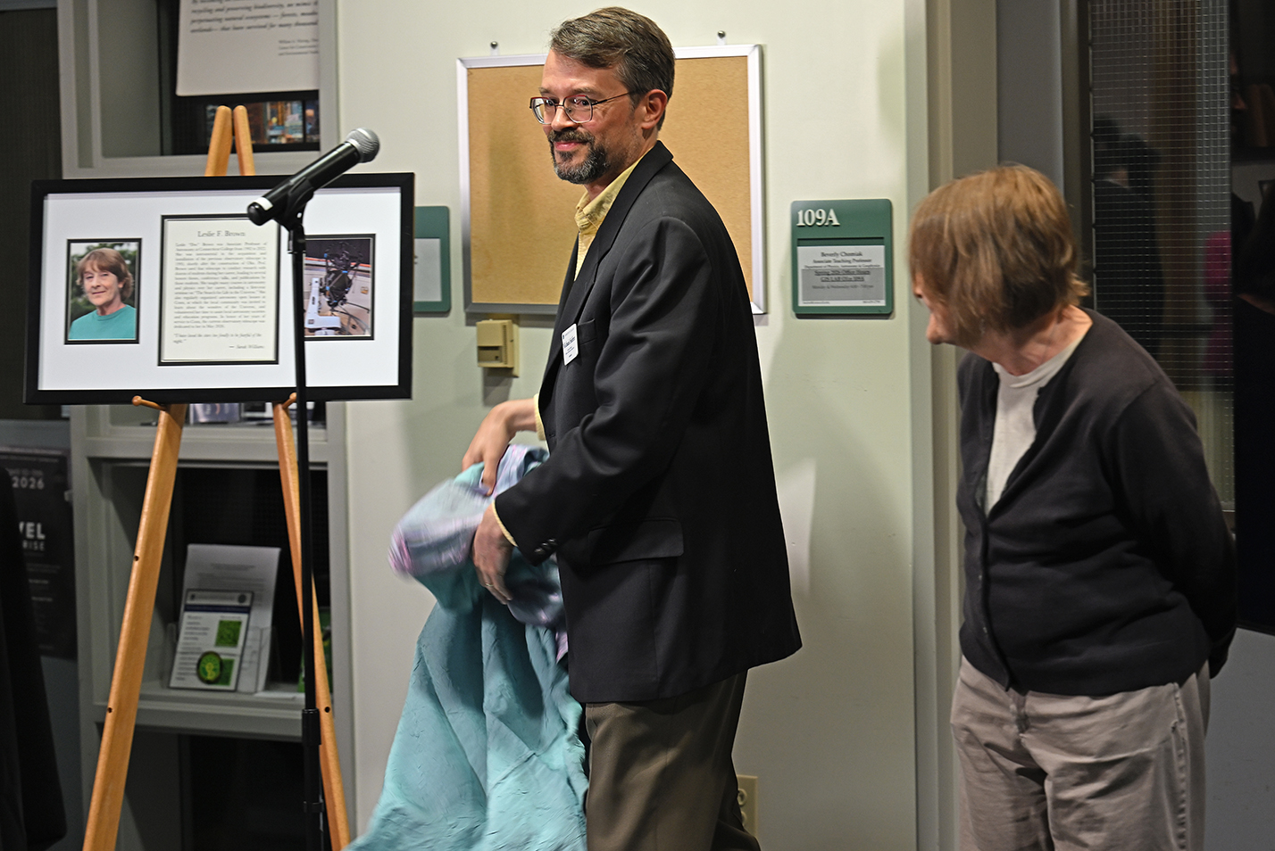 A bearded college professor wearing glasses removes a cover revealing a framed dedication plaque as a retired female professor looks on from the right.