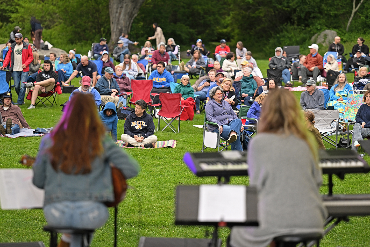 A folk music duo, blurry in the foreground, perform in front of an audience seated on a grassy field.