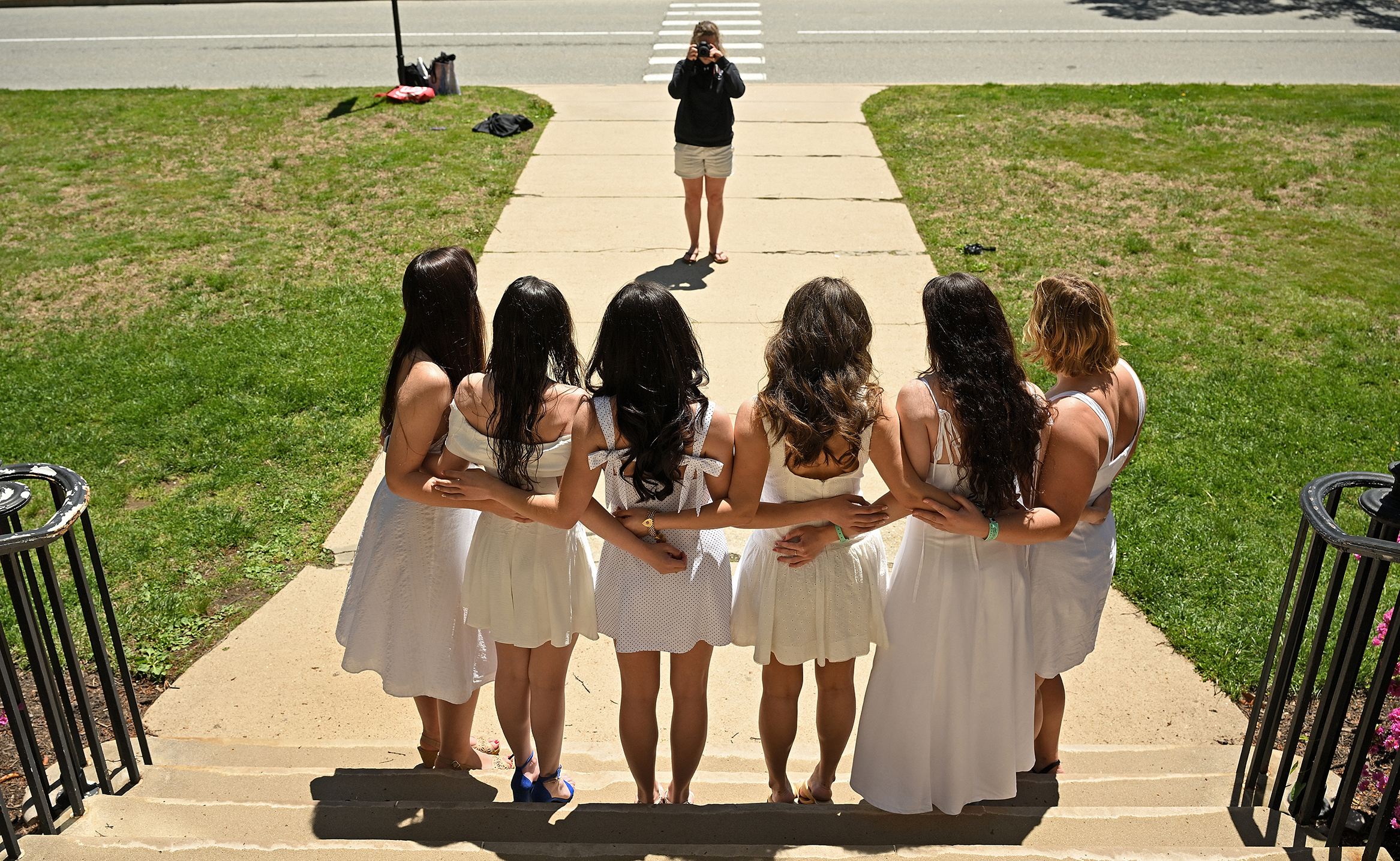 Six college seniors in white dresses facing away from the viewer pose with their arms around each other on the steps of a building for a photographer in the background.
