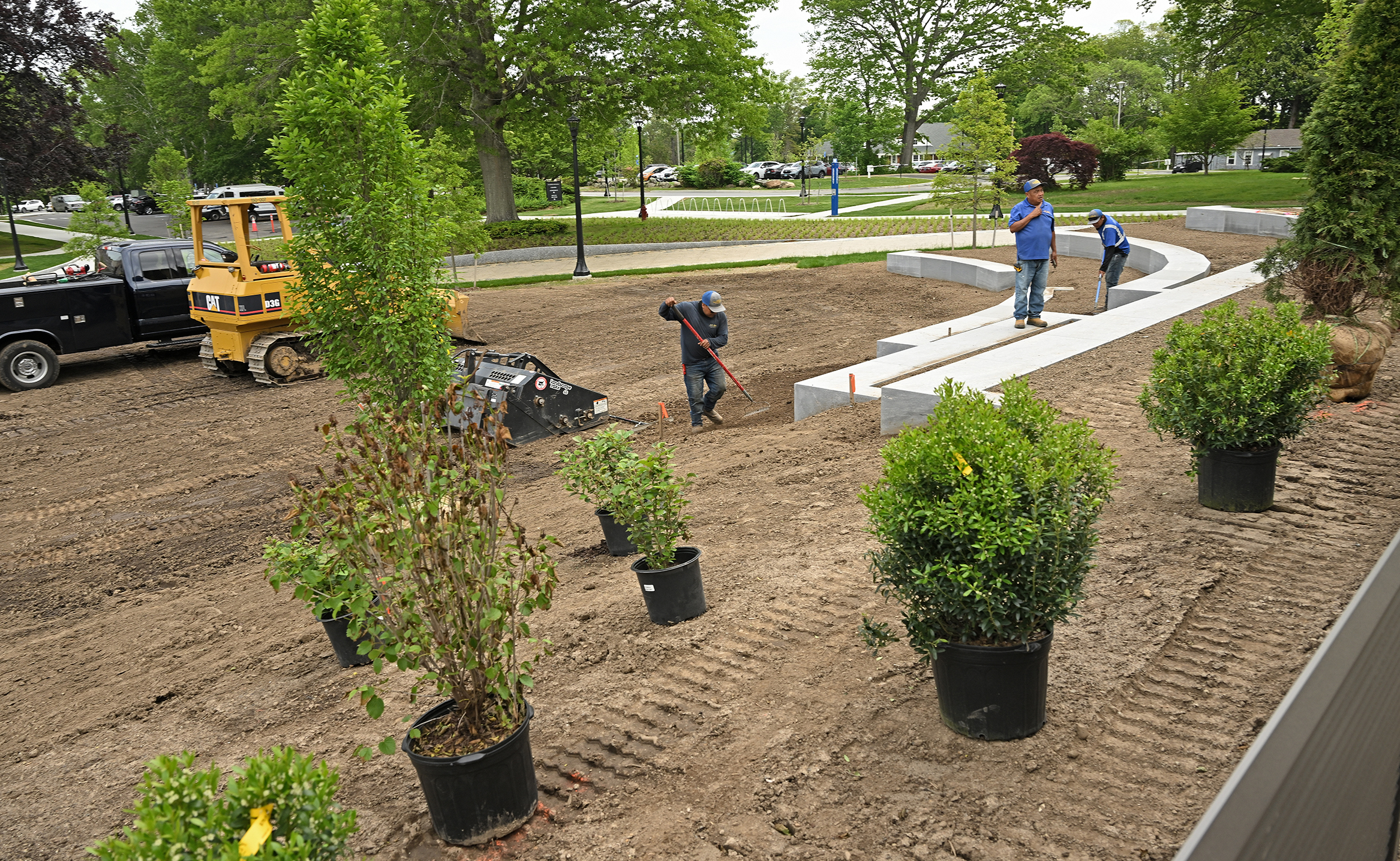 Landscapers work around a dirt area preparing to plant trees and shrubs resting in their pots nearby with a bulldozer and truck in the background.