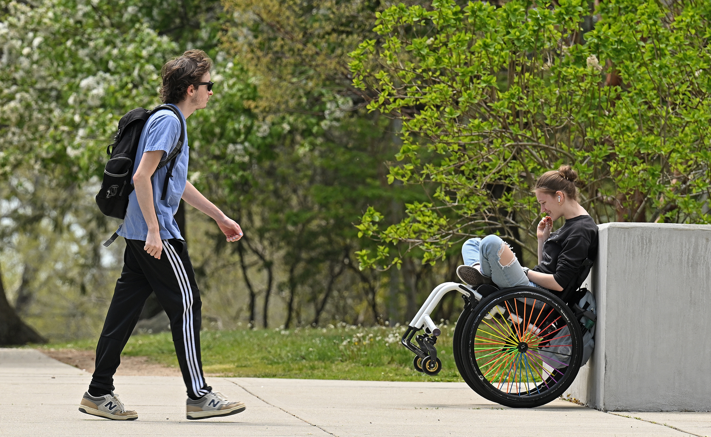 A college student on a wheelchair leans back against a short concrete wall and focuses on the smartphone in her lap as another students walks by.