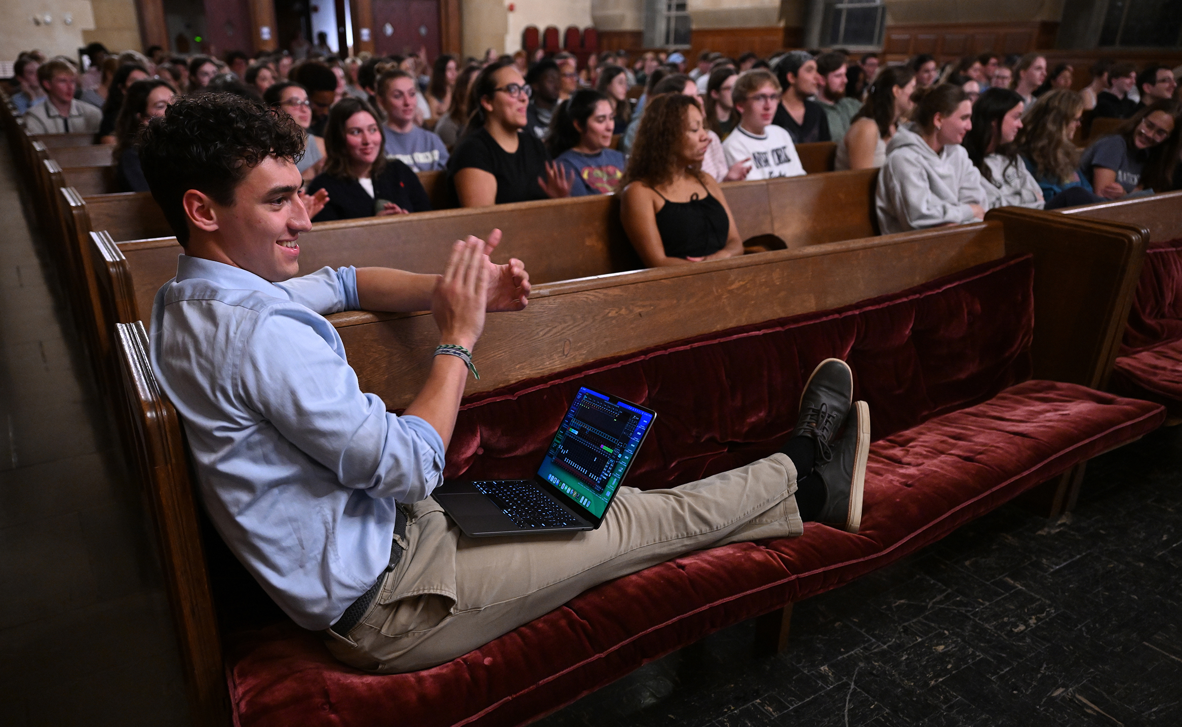 A college students sits with his feet up on the pew with a laptop computer in his lap in a crowded chapel.